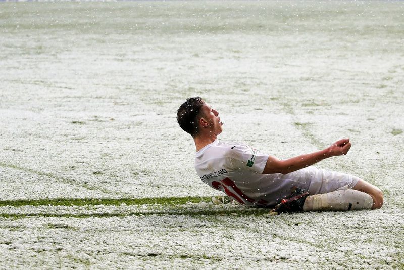 A male football player in a white shirt with red numbering celebrates by sliding on his knees across a snow-sprinkled pitch, leaving a trail of green grass behind him.