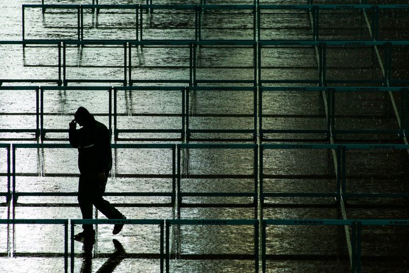 The silhouette of a man holding the hood of his coat to protect himself from heavy rain can be seen walking among rows of hurdles at an athletics ground in this low-light image.