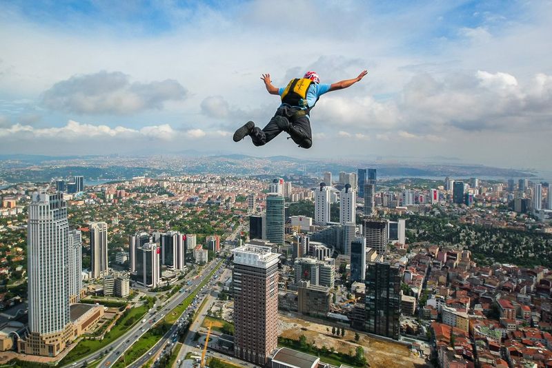 Taken from behind and several thousand feet up in the air, a man wearing a blue T-shirt, black trousers and a parachute skydives above a vast cityscape on a cloudy day.