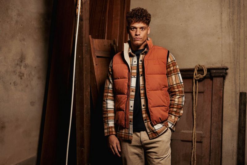 A male fashion model stands against a rustic wooden backdrop wearing a rust coloured gilet over a brown plaid shirt and cream trousers.