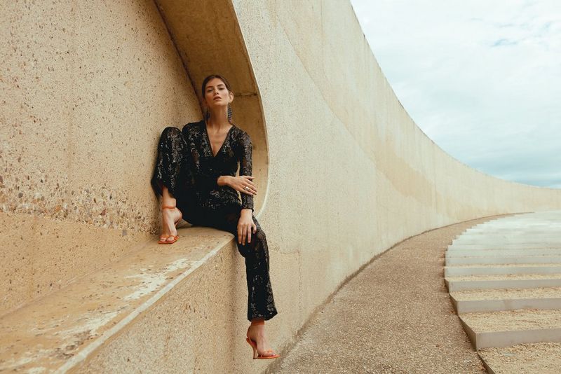 A model wearing a black sequinned jumpsuit and high-heeled orange sandals poses on a ledge inset into a long, curving stone wall. 