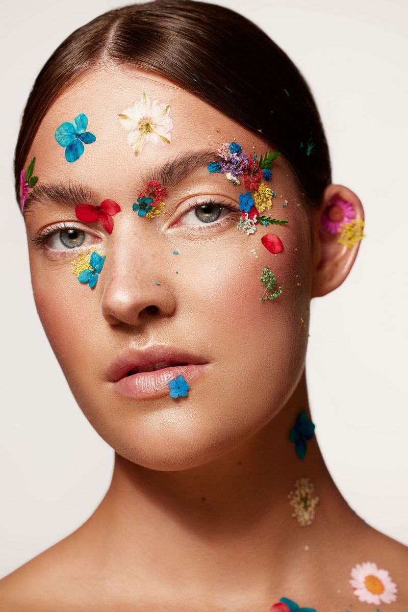 A close-up portrait of a model with her dark hair slicked back and her face decorated with stick-on flowers.