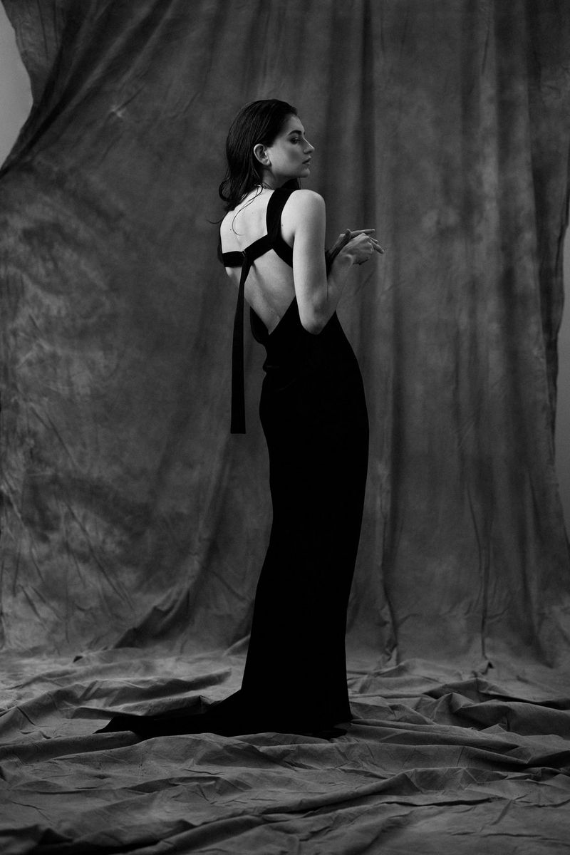 A black and white full-length portrait of a woman wearing a long backless dress.