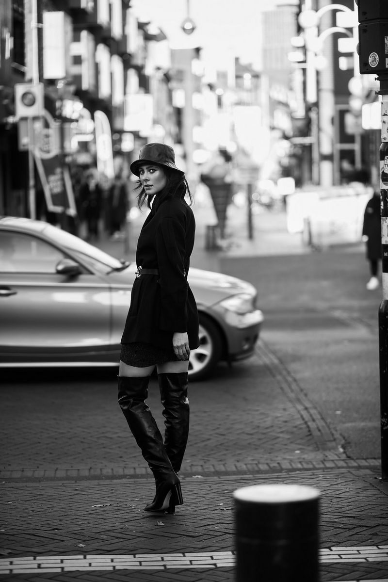 A black a white image of a model wearing a short belted coat and thigh-high boots. She is walking across a busy street while looking back at the camera.