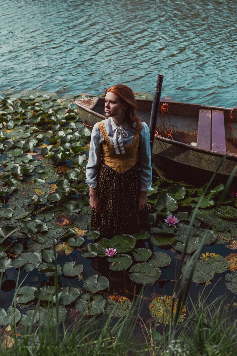 A model standing knee-deep in a lake filled with waterlilies with a wooden rowing boat moored behind her.