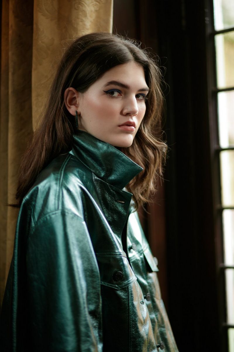 A long-haired model in side profile wearing a shiny dark-green jacket and with her face turned slightly towards the camera. 