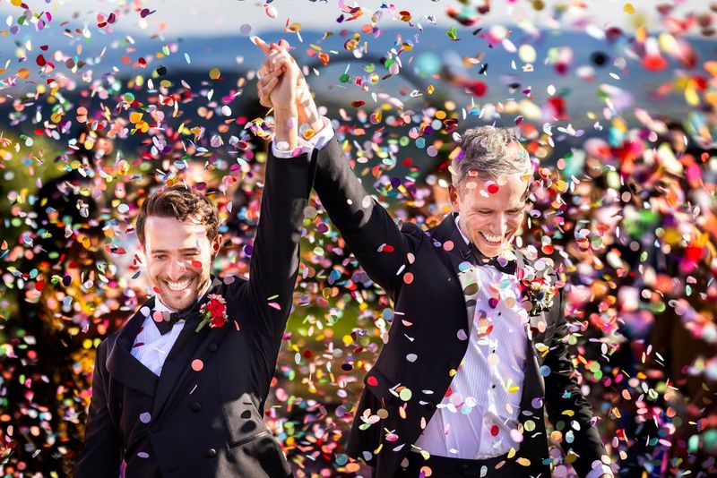 A smiling couple holding hands and lifting their arms in the air while being showered with brightly coloured confetti.