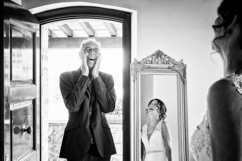 A black and white image of the father of the bride standing in a doorway, smiling with joy. In a full-length mirror you can see the laughing bride's reflection.