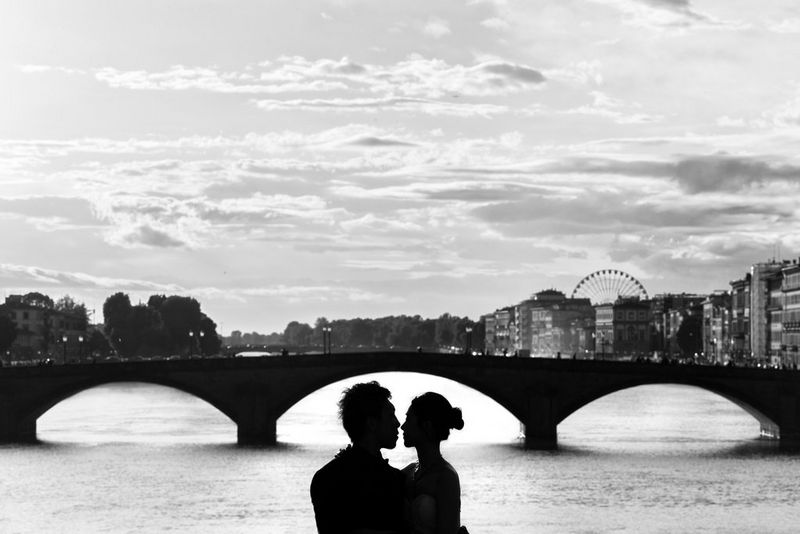 A black and white image of an embracing couple in silhouette, framed by the central arch of a bridge and with a city skyline behind.