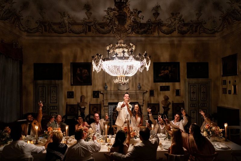 A bride and groom raise a toast to their guests who are seated around a long table under an ornate chandelier. 