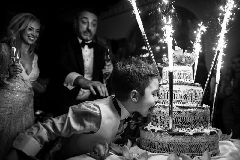 A black and white photograph of a young boy leaning in to take a bite out of a tiered wedding cake, the groom behind reaching to stop him. 