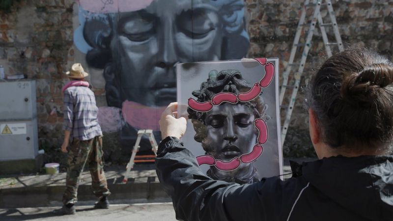 In the foreground, a man holds up an image of a statue's head covered in strings of pink cartoon sausages. In the blurred background, a second man paints sausages across a large image of the same head on a wall. Still from Best Before by Filippo Tommasoli.