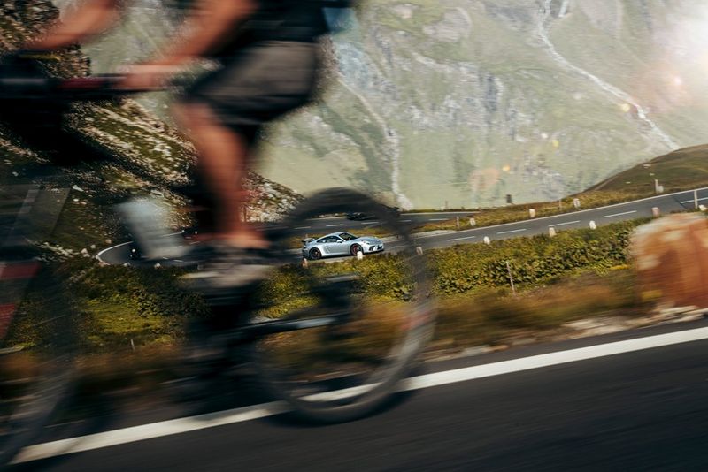 A Porsche driving up an alpine road framed by the rear wheel of a bicycle, taken on a Canon EOS R by Florian Roser.