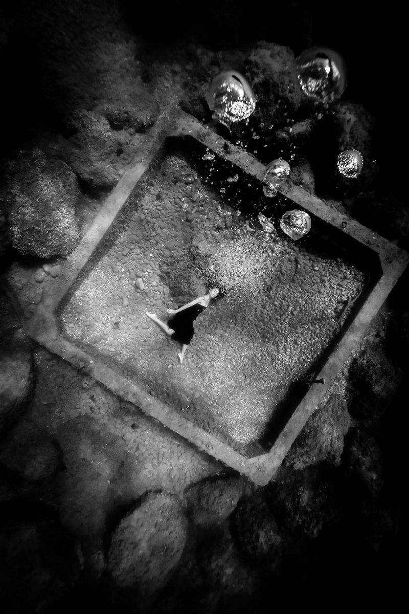 Une image en noir et blanc d'une femme portant une robe, allongée sur le fond marin au centre d'un cadre géant. Une ribambelle de bulles s'élève, flottant vers l'appareil photo.