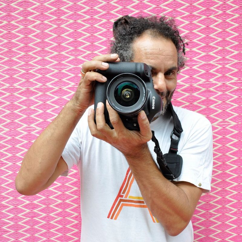 Canon Ambassador Hassan Hajjaj holding a Canon camera, against a pink background with a vertical zig-zag pattern. 
