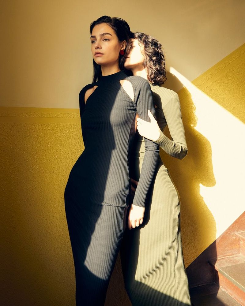 Two models in tight-fitting dresses, one holding the other's arms from behind, stand against a yellow wall, lit by a shaft of light. Taken by Canon Ambassador Heidi Rondak