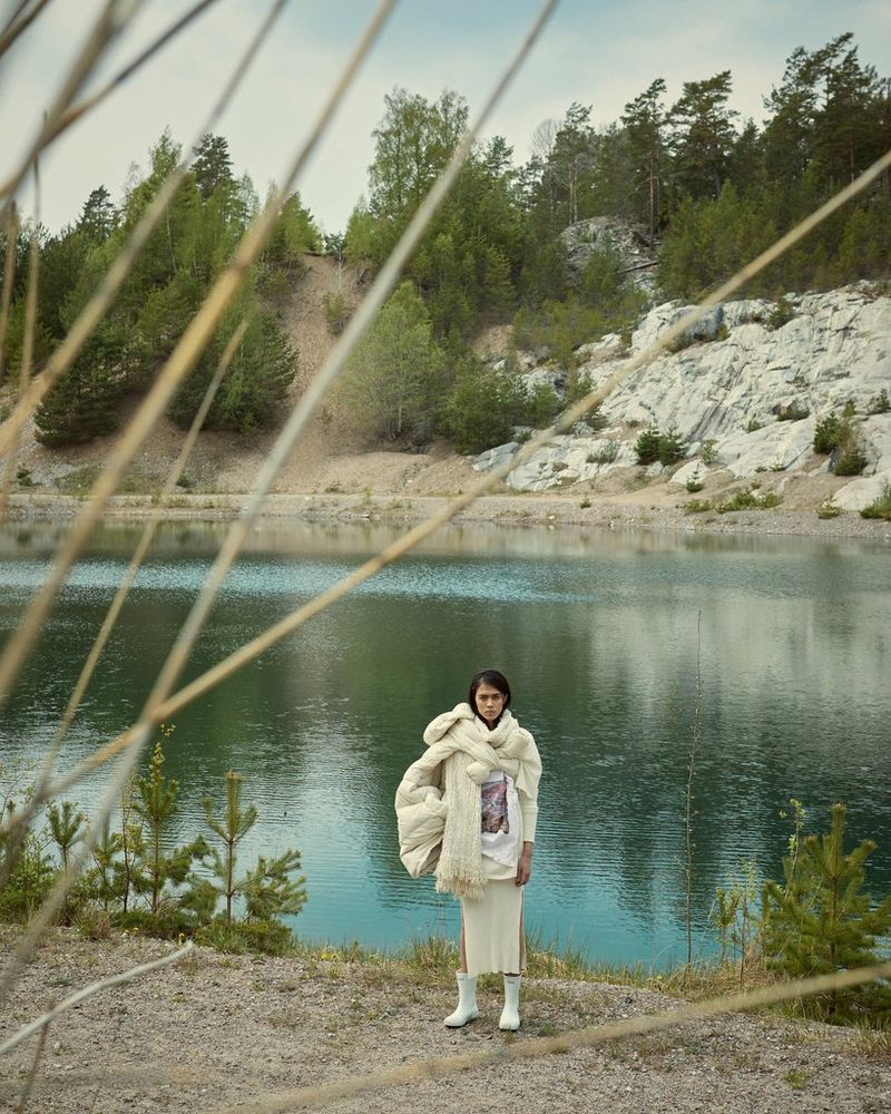 A model stands in front of a lake, wearing white boots, a cream knitted skirt and with a cream quilted jacket and an oversized scarf around her neck, in a portrait by Canon Ambassador Heidi Rondak.