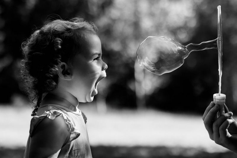 A black and white photo of a young girl with her mouth open wide in wonder as she watches the bubble in front of her face about to burst.