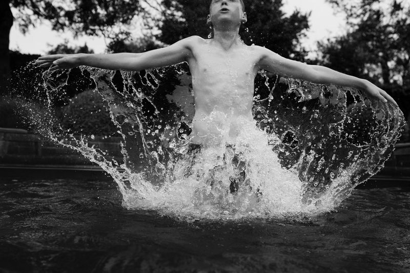 A black and white image of a boy jumping out of a swimming pool, splashing water all around him.