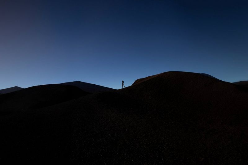 Une coureuse traversant des dunes volcaniques, sa silhouette et celles des dunes se découpant sur le ciel bleu foncé.