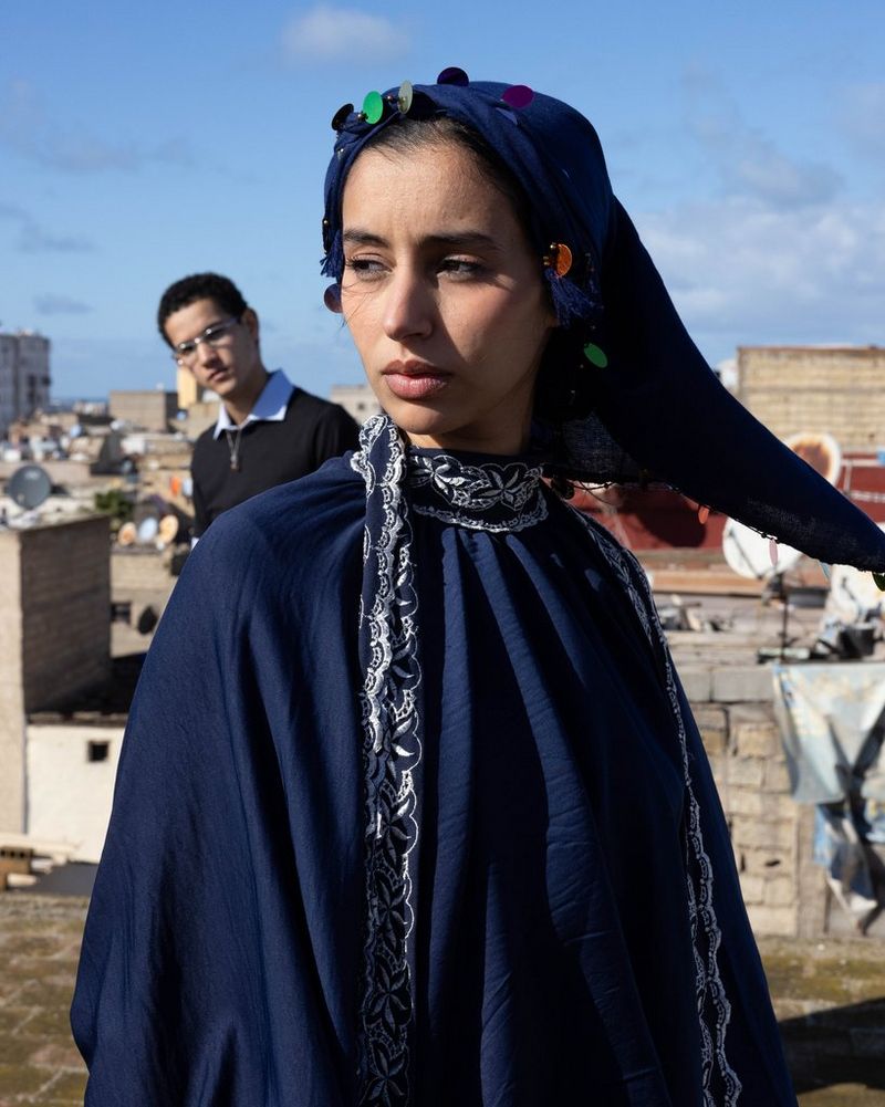 Une femme, portant un foulard et une robe bleue, debout sur un toit, avec un homme juste derrière elle.