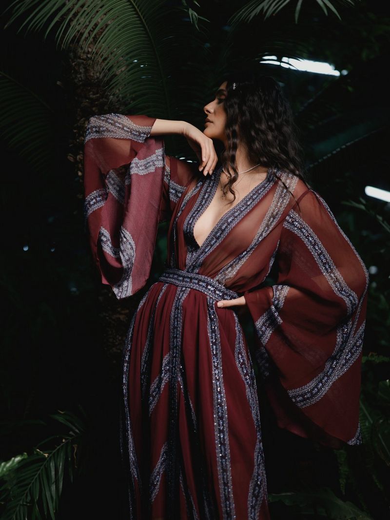 A model wearing a burgundy kaftan posing against a backdrop of green foliage.
