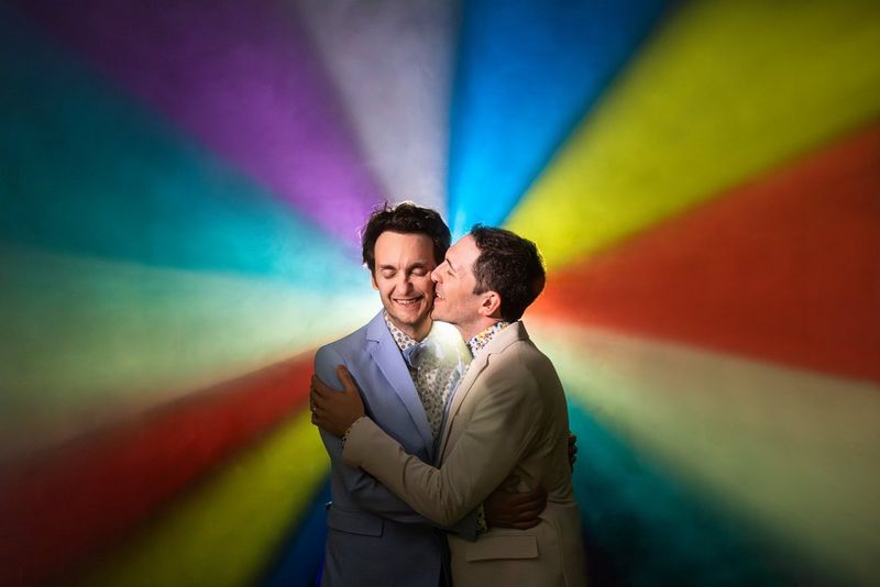A medium close up of two men at their wedding, embracing and kissing in front of a colourful lit-up background. Taken on an EOS R5 Mark II by Canon Ambassador Fabio Mirulla.