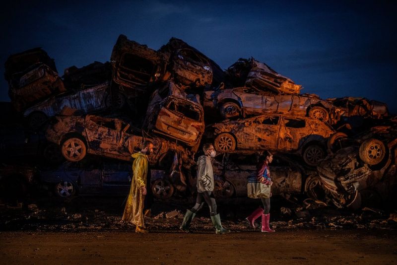 A low light image of three people walking in front of a pile of stacked, damaged cars, covered in mud. Taken on the EOS 5D Mark IV by Canon Ambassador Santi Palacios