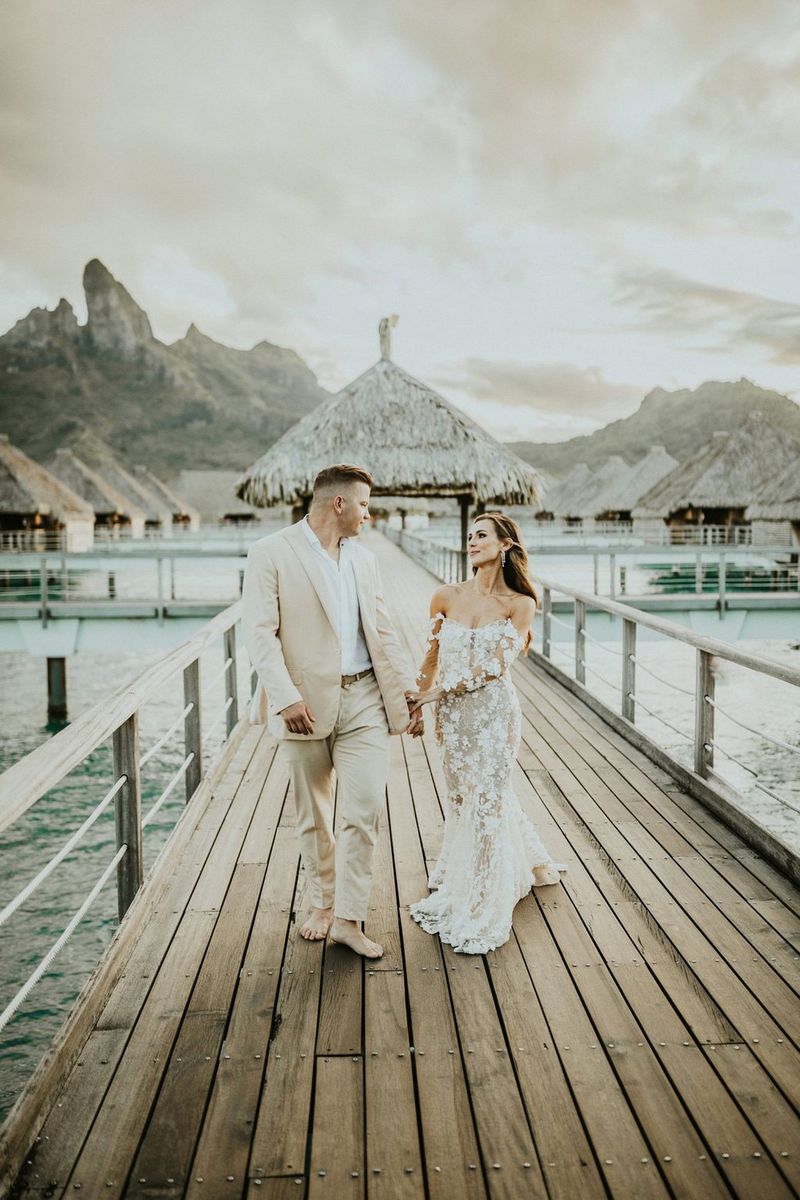 A newlywed couple walk hand in hand down a wooden jetty, thatched overwater bungalows in the background. Taken by Igor Demba.