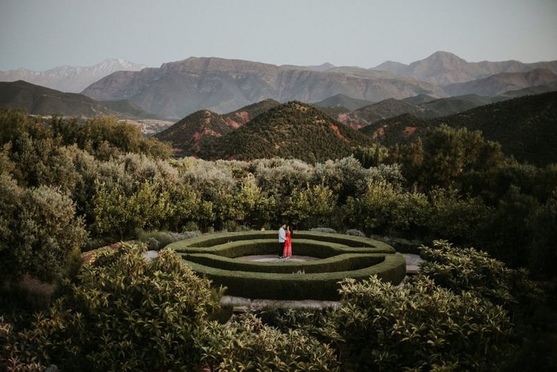 A couple standing in the centre of a circular parterre, with trees and mountains in the background. Taken by Igor Demba. 