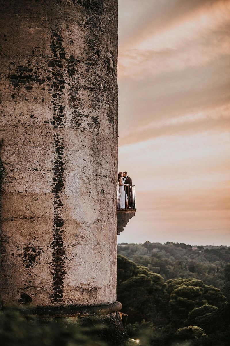 A couple standing on a balcony with wrought iron railings on the side of a large stone tower. Taken by Igor Demba.