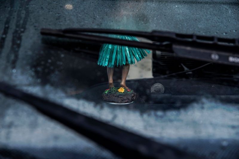 Through the gap in the wipers on a car's windscreen, the legs of a hula dancer doll are visible. The doll is stuck on the dashboard; next to it is a coin. Taken by Iiris Sjöblad.