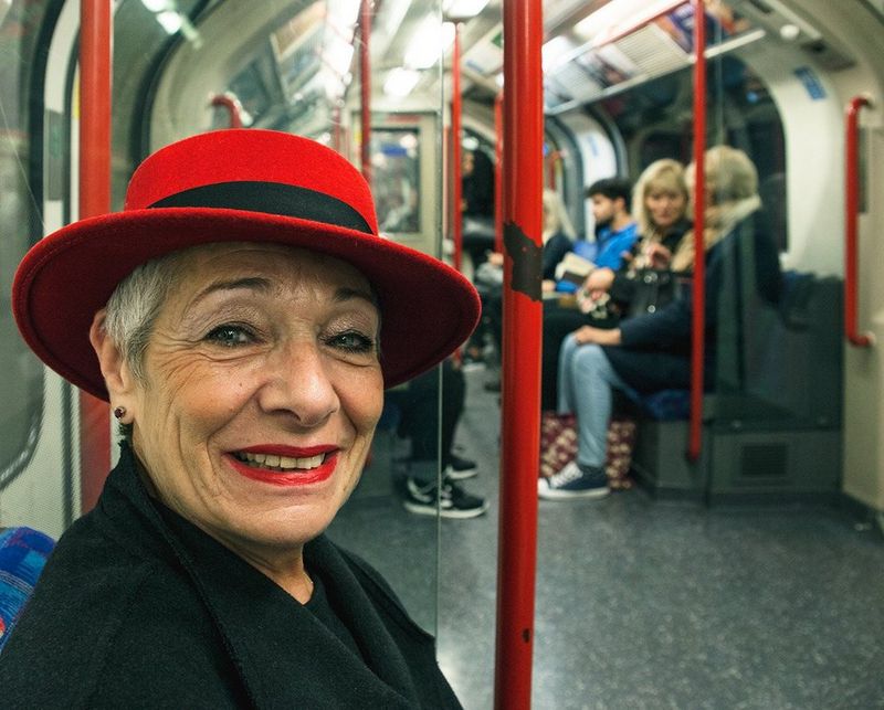 A woman on the London underground wears a red hat and smiles at the camera. Her hat matches the red railings behind her. Taken by Iiris Sjöblad. 