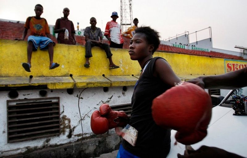 A young black woman is helped into boxing gloves by her coach, while a group of young men watch on.