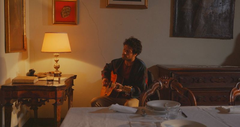 A man strumming a guitar sits behind a table inside a room lit by low light. 