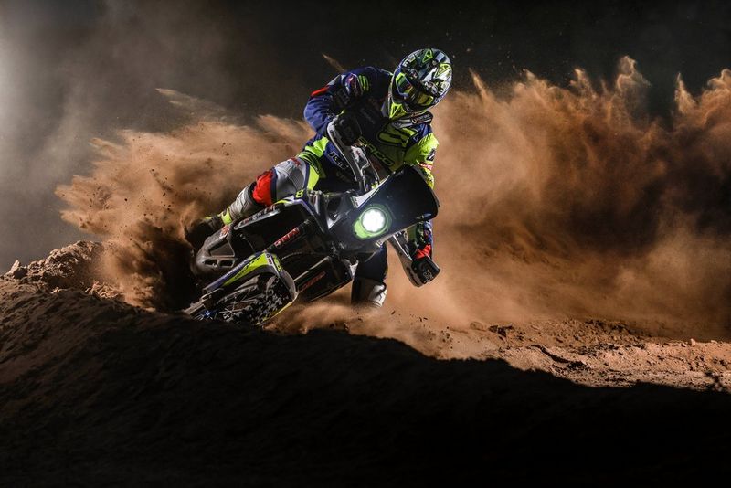 A motorcyclist driving round a corner on a muddy track, kicking up a huge cloud of dirt.