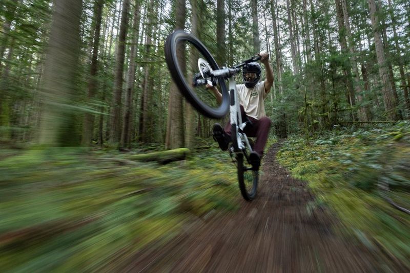 A mountain bike rider performs a wheelie along a forest trail, the foreground blurred by a long exposure.