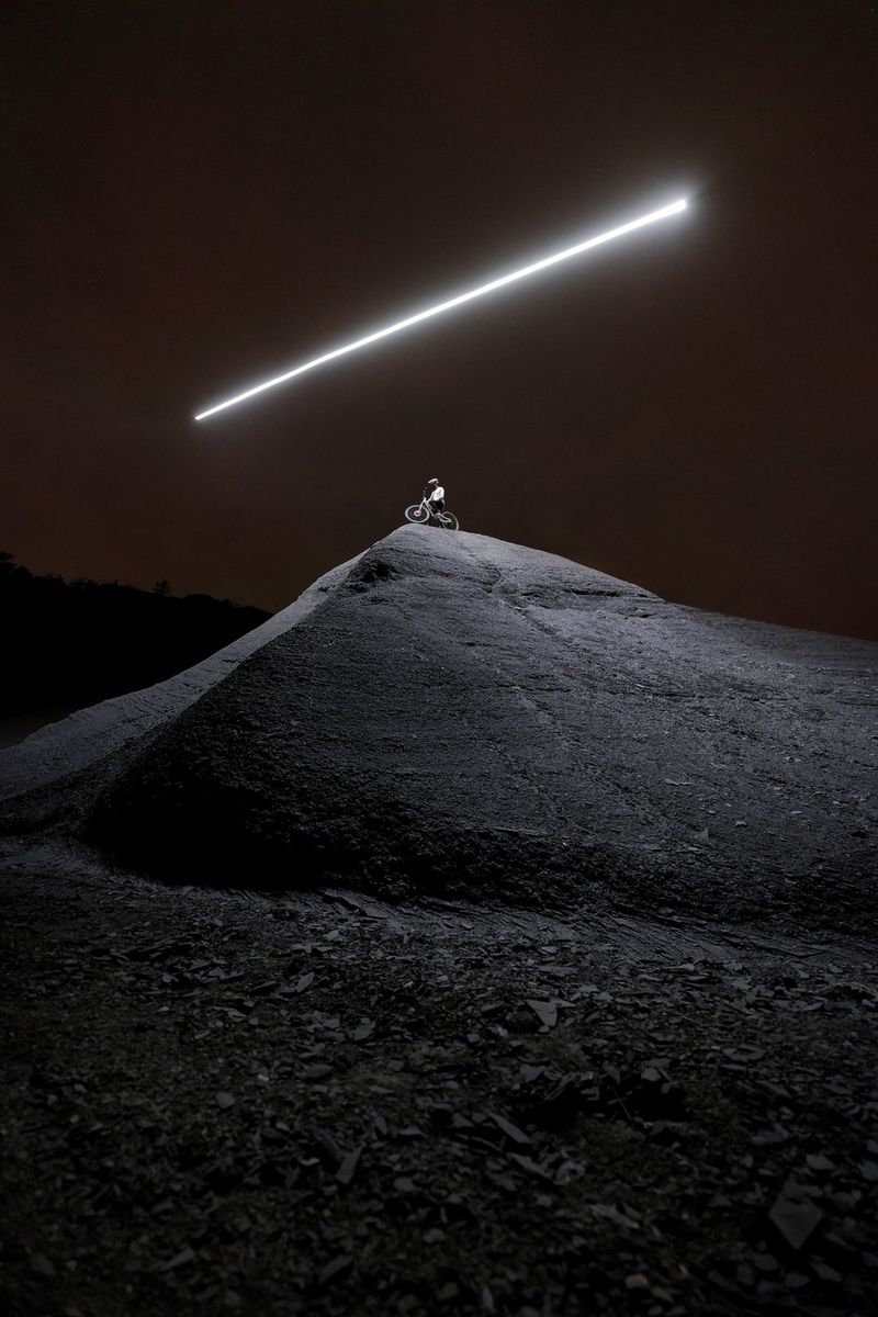 A nighttime long exposure of a mountain bike rider atop a hill, with a single beam of light in the dark sky above.