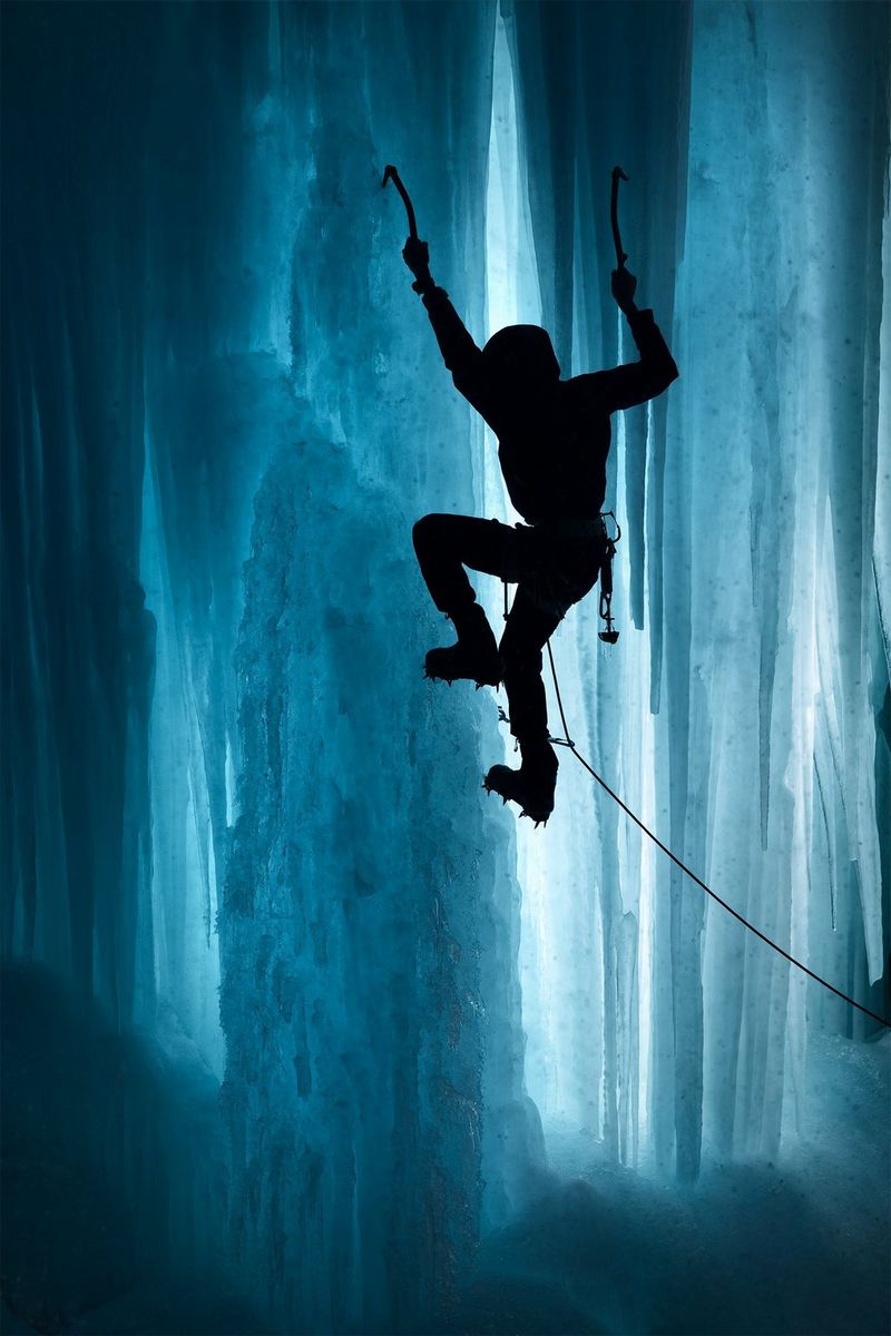 An ice climber silhouetted against the blue wall of ice he is ascending.
