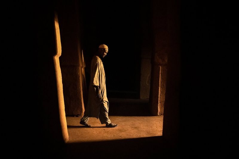 A low-light, minimalist portrait of a man leaving a mosque framed by dark shadows.