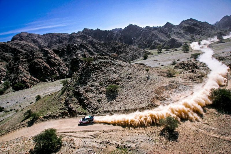 A rally car leaves behind a huge trail of dust as it races around a desert track.