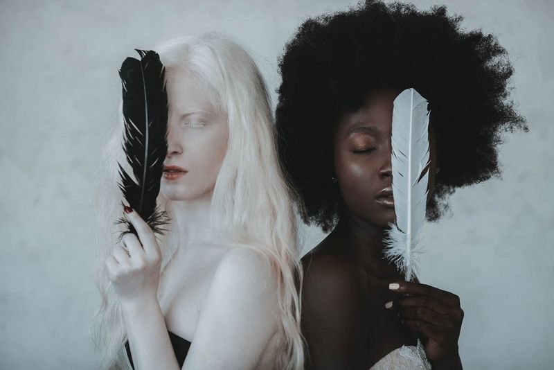 A white albino woman with blonde hair holding a black feather and a black woman with an afro holding a white feather. Both women have their eyes closed and their backs to each other.
