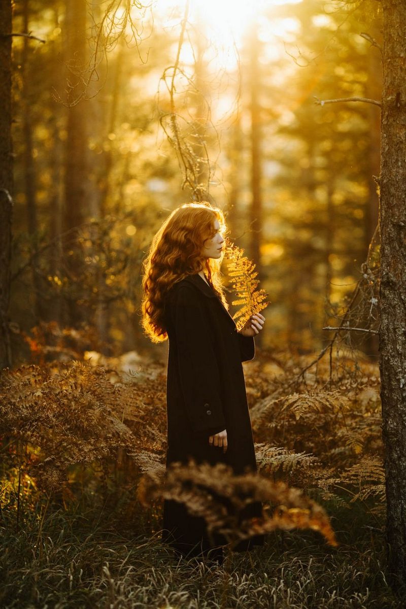A side profile portrait of a model with red hair bathed in orange light, standing in a forest amidst a sea of faded ferns and holding a brown fern leaf.