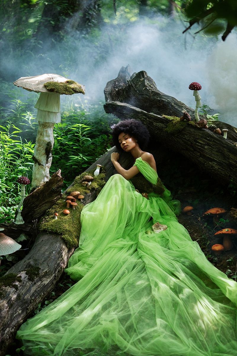 A model in a bright green dress reclining on a moss-covered fallen tree trunk in the shade of a giant mushroom.