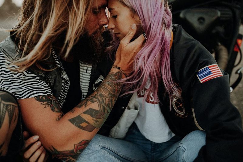 A blond man with tattoos on his forearms embraces a woman with lilac hair.