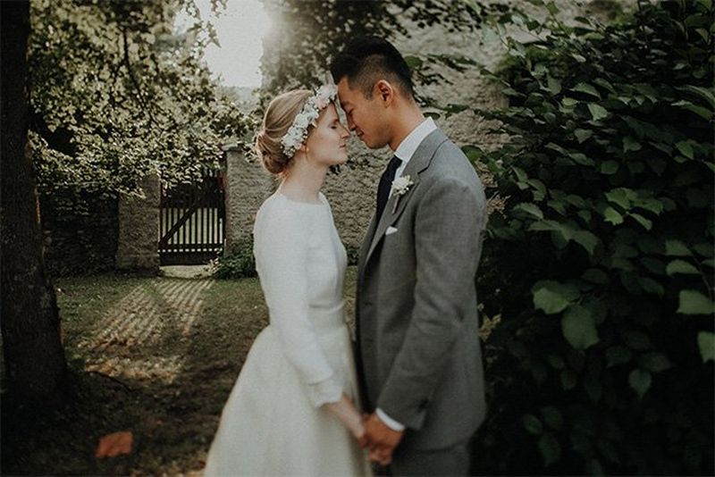 A bride and groom stand face to face in a leafy garden; they are holding hands and pressing their noses together. 