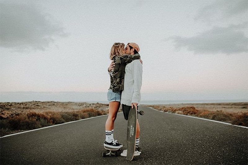 A woman kisses a man as he holds a skateboard in the middle of an empty road.