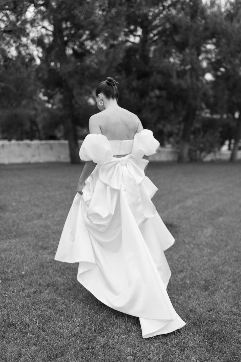 A bride in a backless dress with puff sleeves and a large bow walks away from the camera, holding her skirt up off the grass.