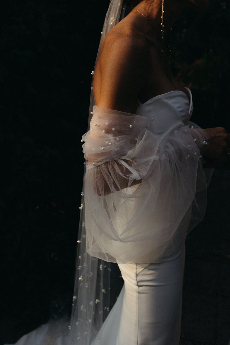 A side profile of a bride, shot from the neck down, wearing a figure-hugging strapless dress and a veil studded with pearls.
