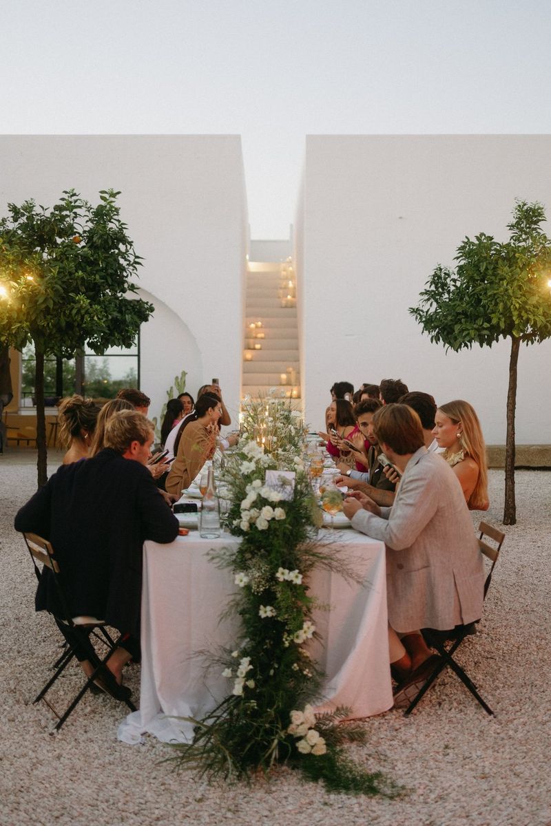 A wedding party sits at a long outdoor table with a large floral centrepiece.